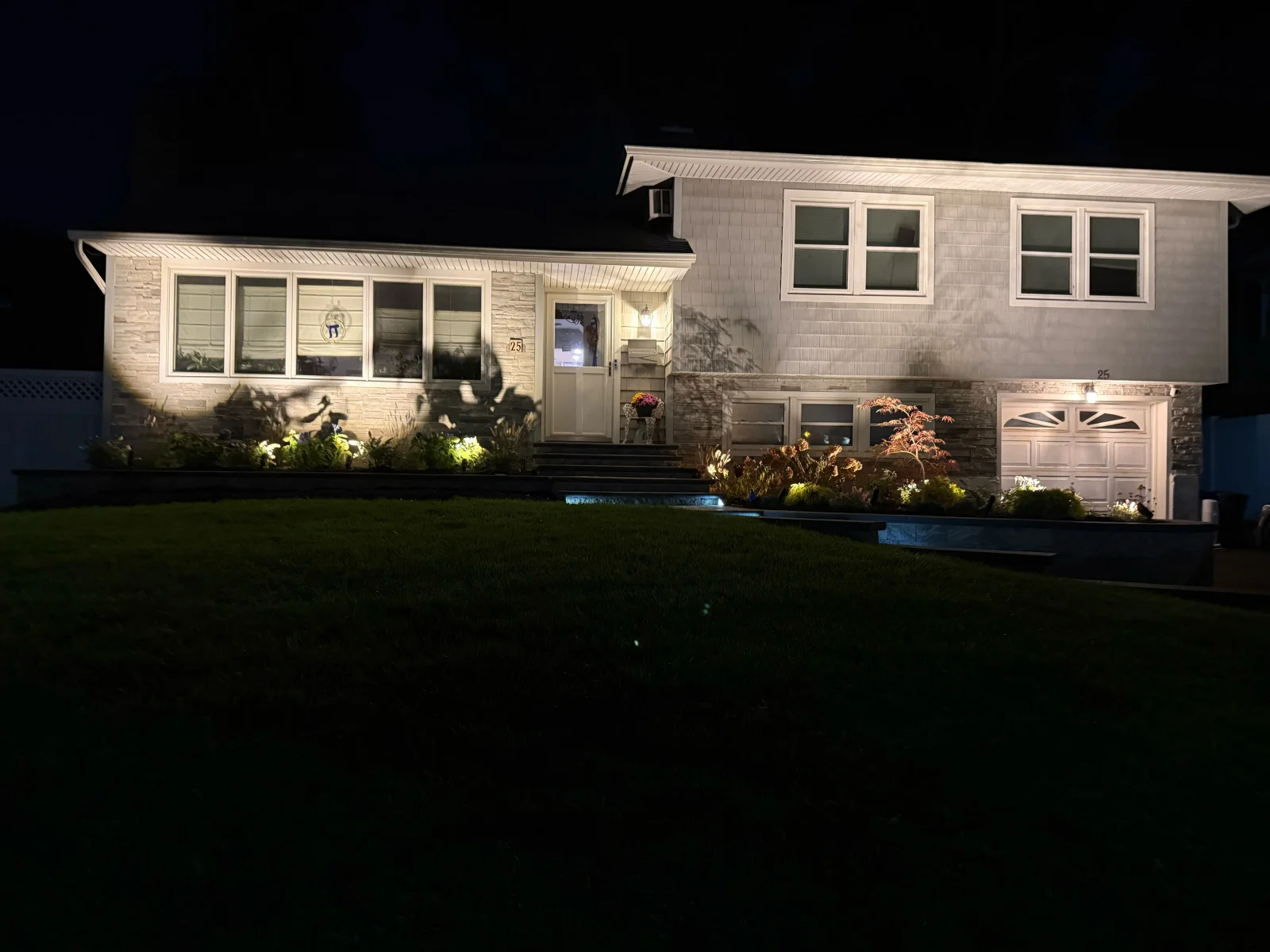 Night view of split-level home with uplit stone veneer, planting beds, Japanese maple, and retaining wall lighting