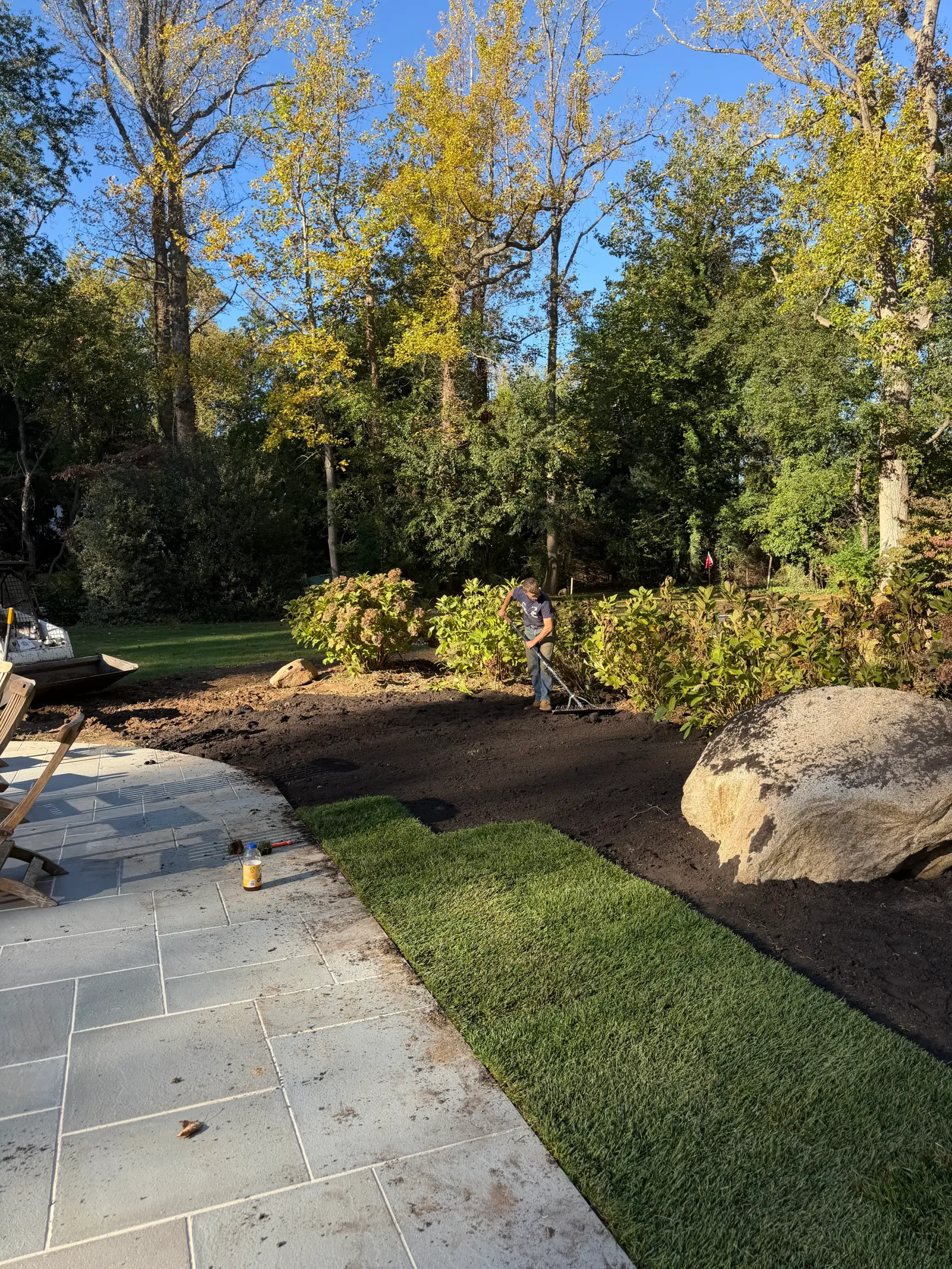 Worker raking dark topsoil beside rolled sod, boulder, and shrubs next to a gray flagstone patio