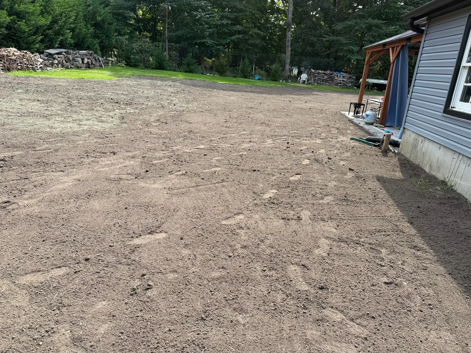 Freshly graded brown topsoil across a backyard beside a gray vinyl-sided house, prepared for seeding or sod installation.