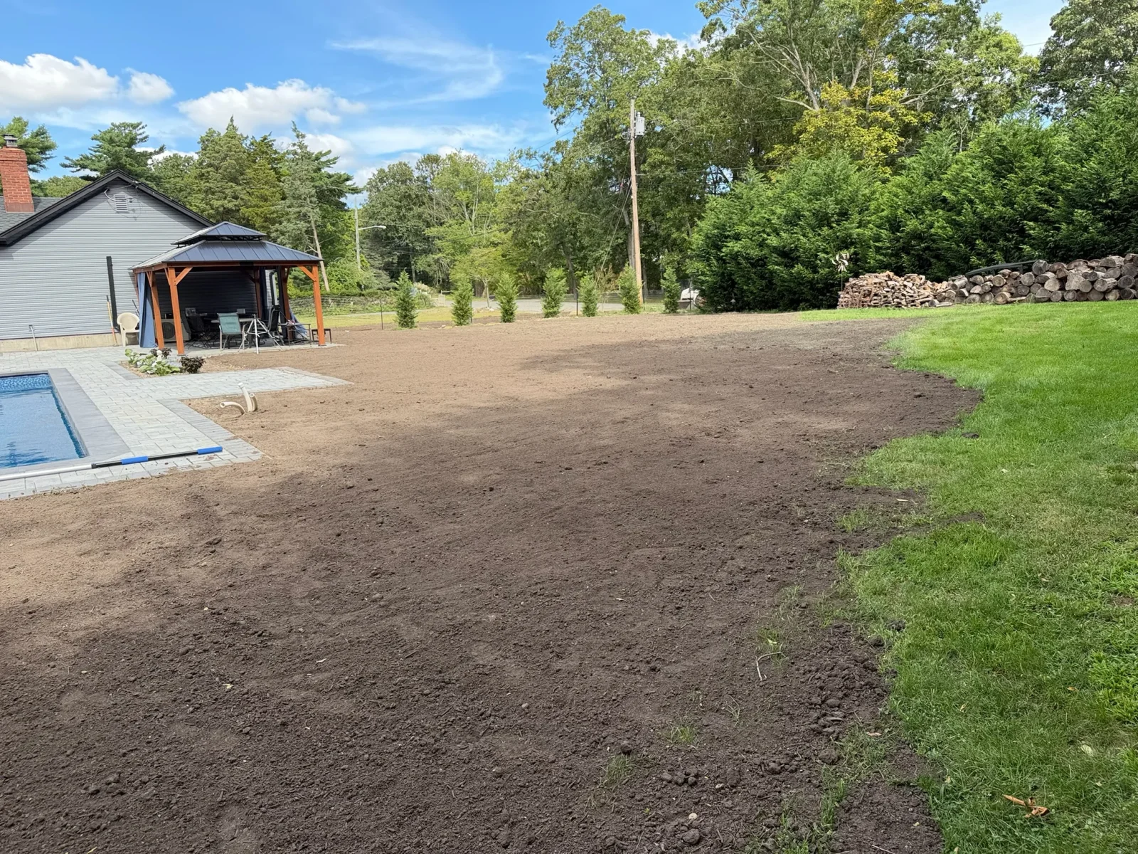 Freshly graded dark topsoil in a backyard beside gray paver patio, in-ground pool, and wooden gazebo with metal roof