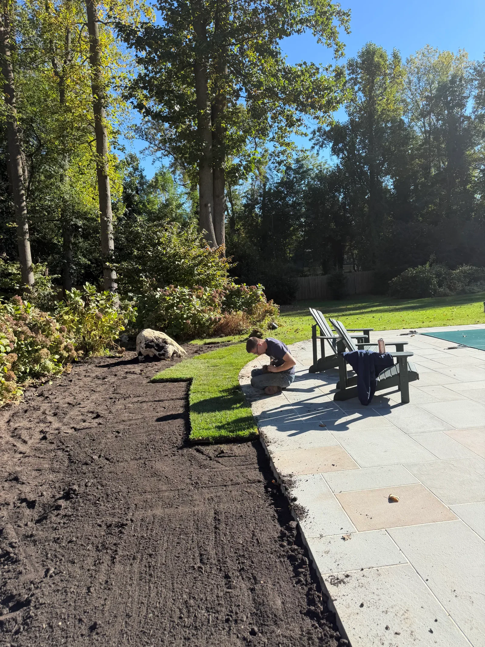 Worker laying sod beside gray stone patio with bare soil prepared for lawn, boulder and shrubs nearby