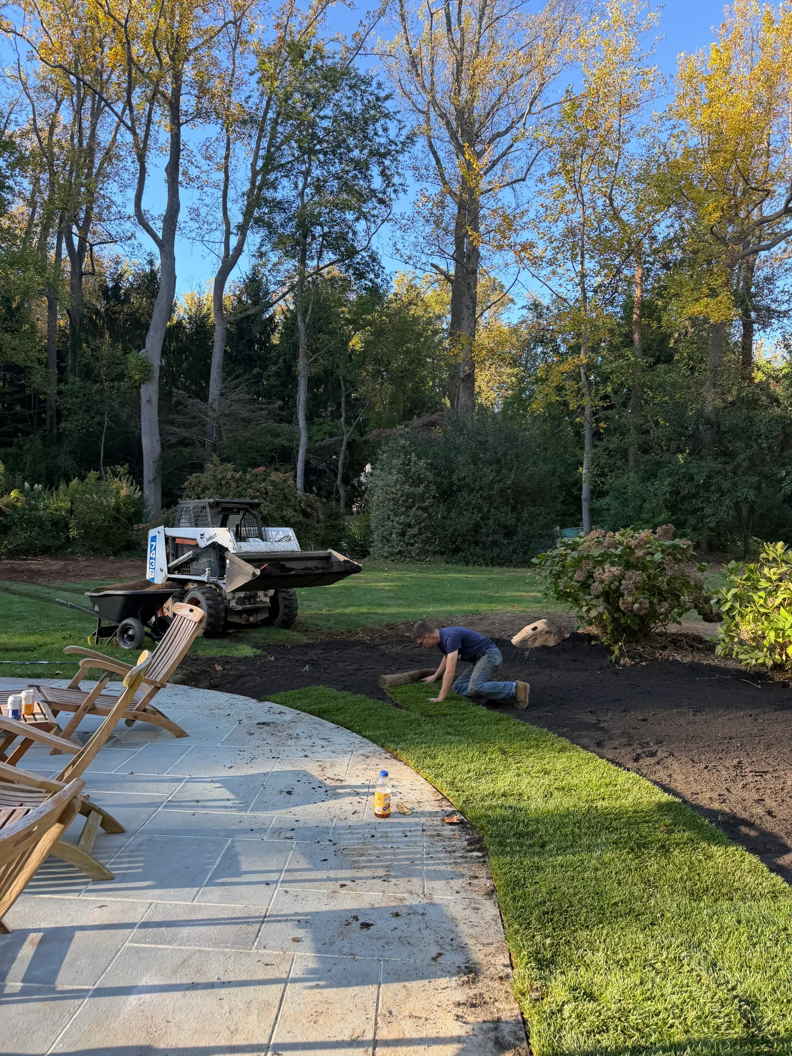 Worker laying sod on graded topsoil beside a gray stone patio, with a Bobcat skid steer grading the lawn