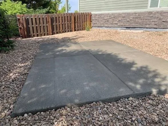 Gray concrete pad with control joints surrounded by gravel and river rock, beside a wood privacy fence and stacked stone veneer wall