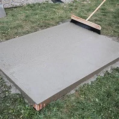 Freshly poured concrete slab with broom-finished texture, wooden form boards, and push broom on grass