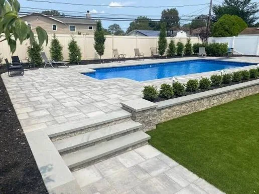 Gray paver pool patio with stone steps, stacked stone retaining wall, low shrub plantings, and artificial turf lawn beside an in-ground pool