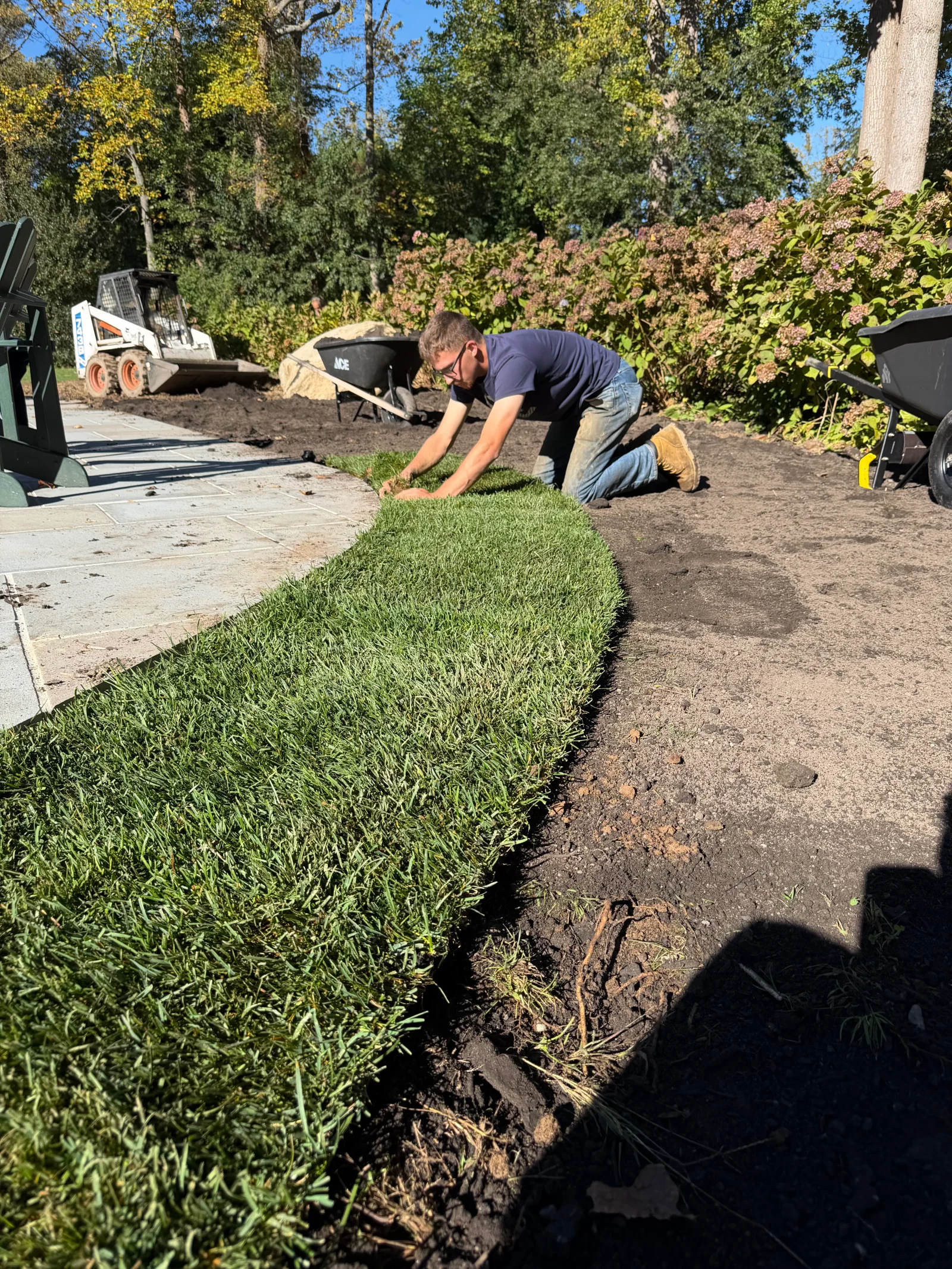 Worker laying sod on graded soil beside a gray flagstone patio, with a skid steer and wheelbarrows nearby