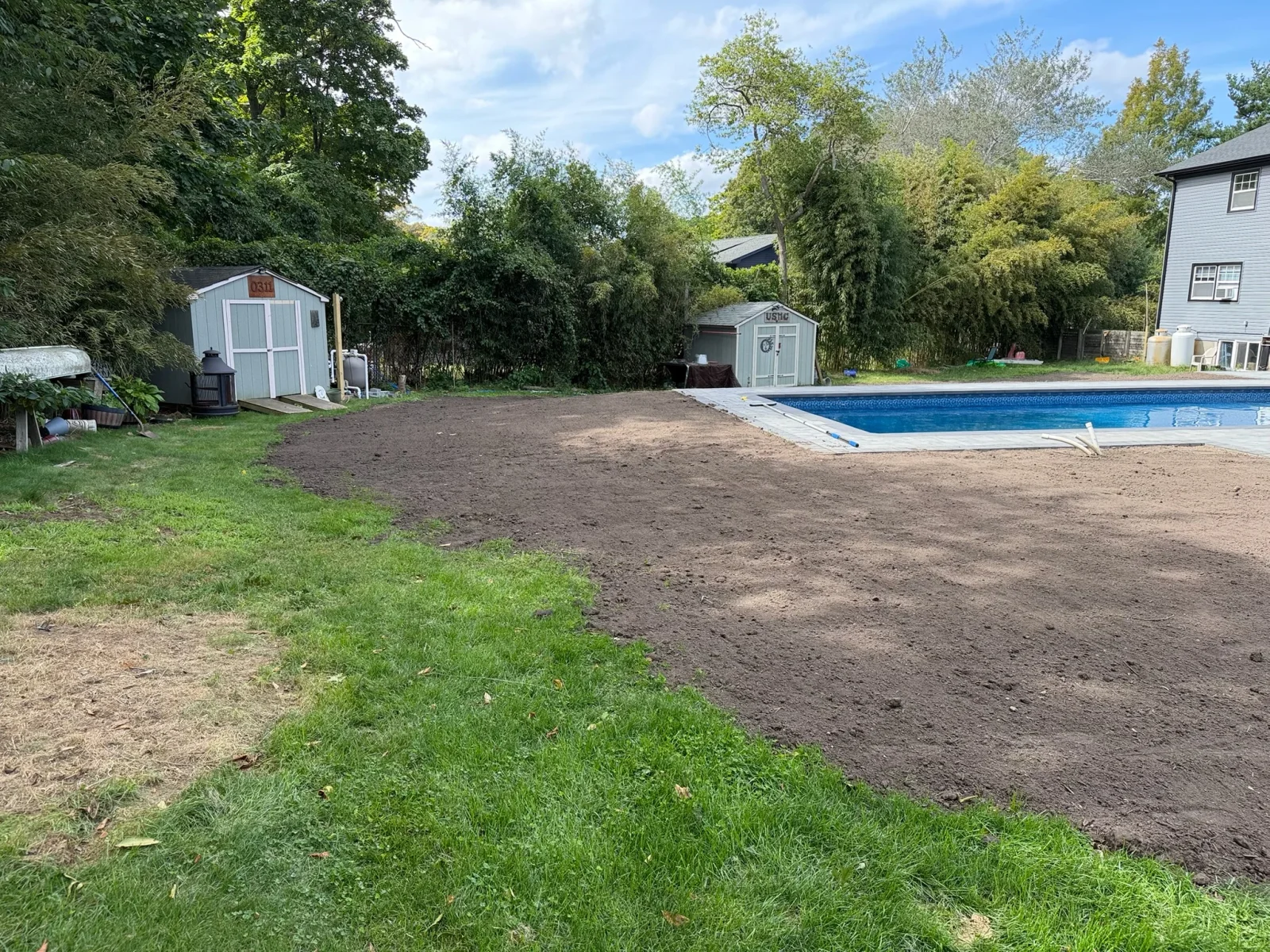 Freshly graded dark topsoil beside an in-ground pool with gray paver coping, next to established lawn and garden shed