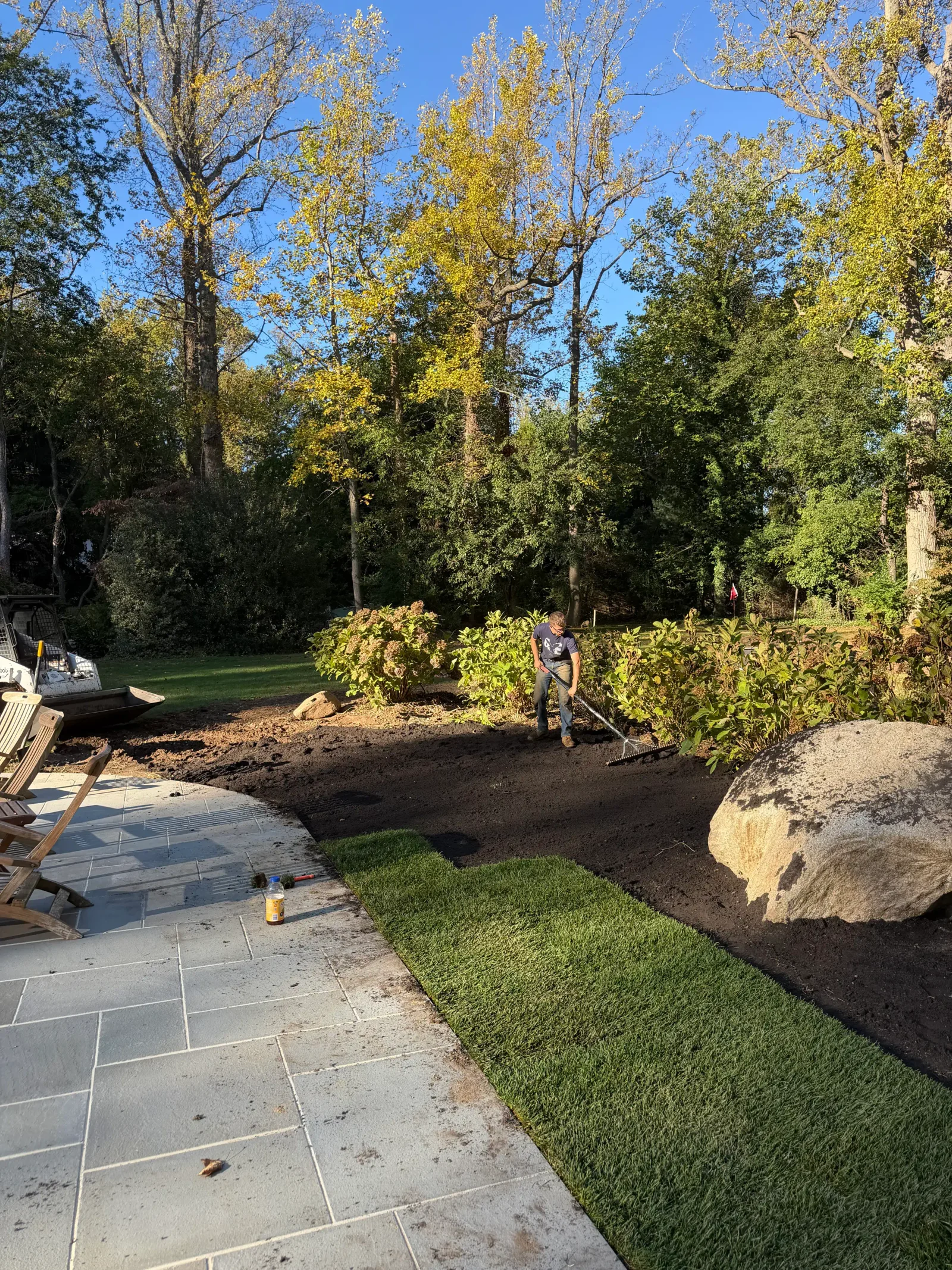 Worker raking topsoil beside freshly laid sod and gray flagstone patio with boulders and shrubs