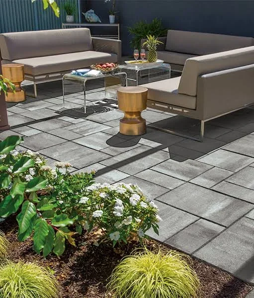Large gray pavers on a patio with taupe sectional sofas, glass tables, and white flowering shrubs in mulched beds