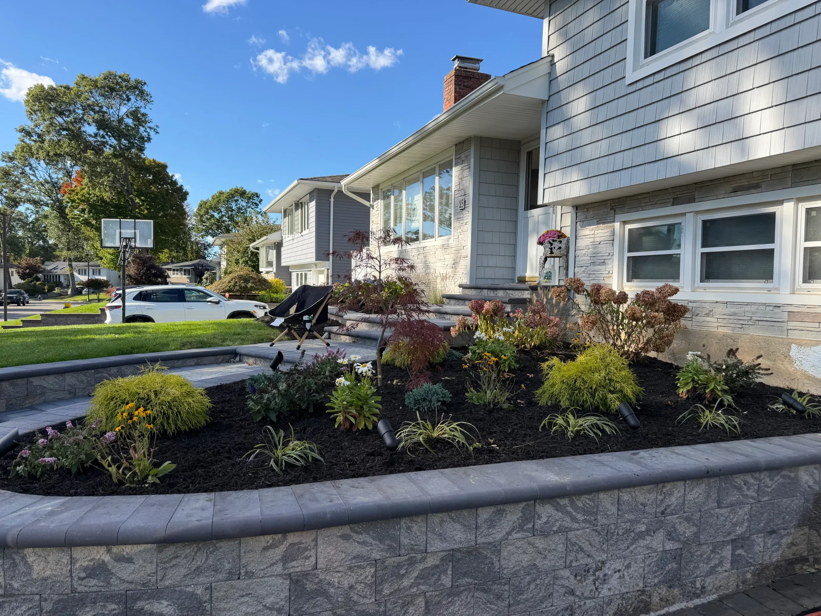 Front yard planting bed with gray segmental retaining wall, dark mulch, ornamental grasses, Japanese maple, and perennials